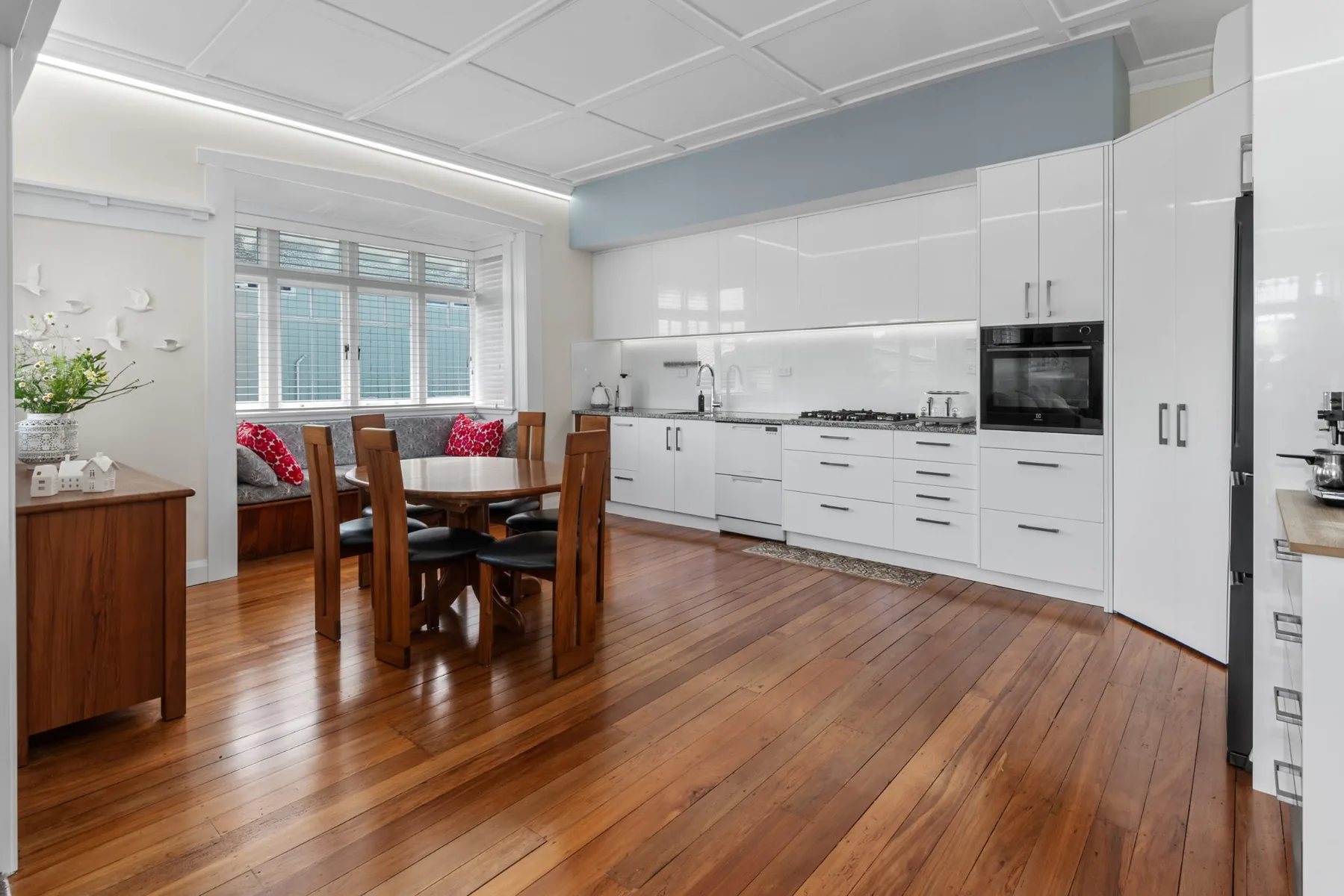 Relocated white kitchen beside the dining area, with timber flooring and built-in appliances